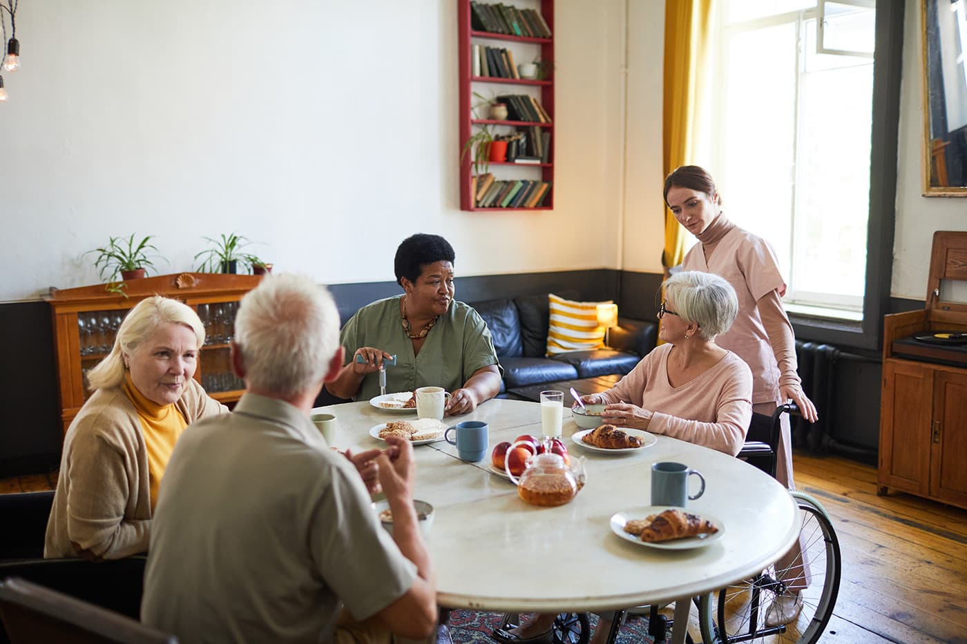A caregiver stands beside a diverse group of older adults gathered around a dining table in a residential care setting.