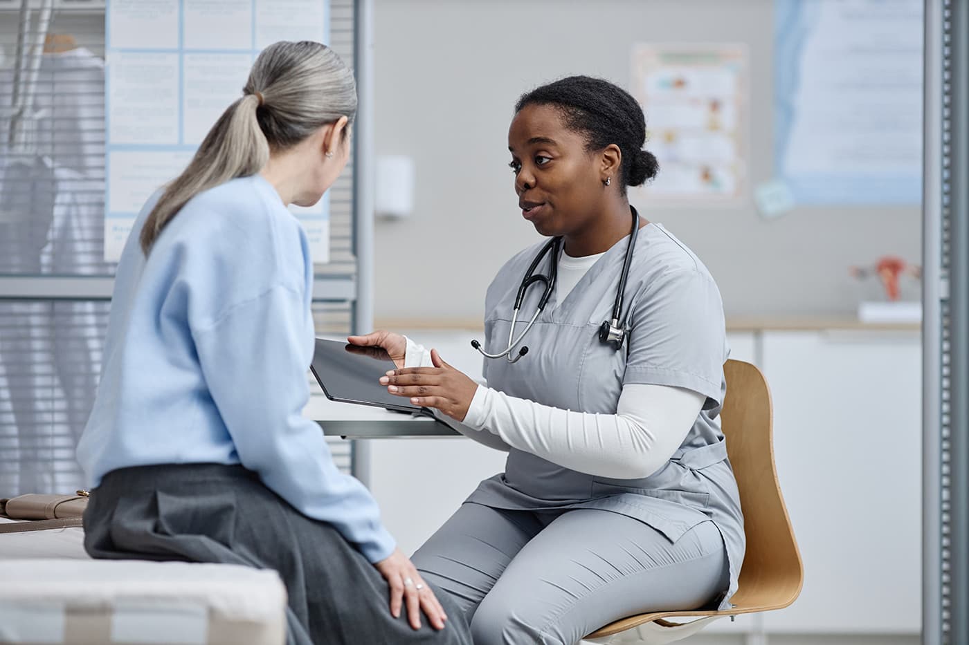 A nurse in gray scrubs shows a tablet screen to a patient during a seated consultation in a clinical setting.
