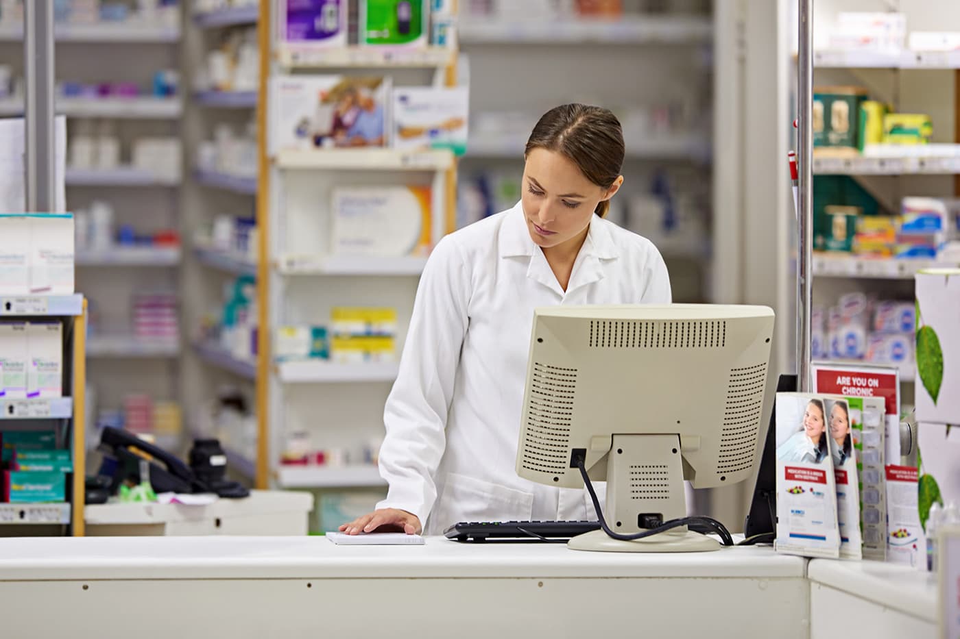 A pharmacist in a white lab coat reviewing information on a computer at a pharmacy counter